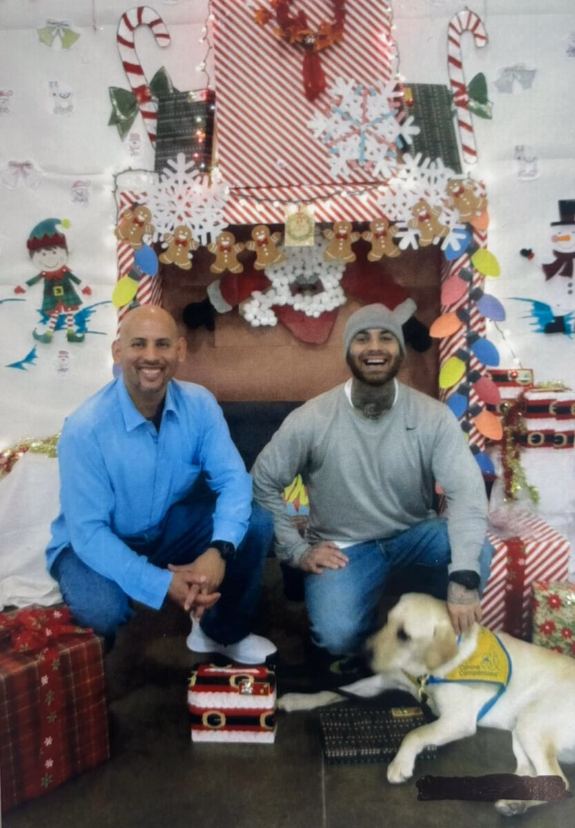 Two men and a yellow Labrador retriever wearing a service dog vest pose in front of a decorated Christmas fireplace.