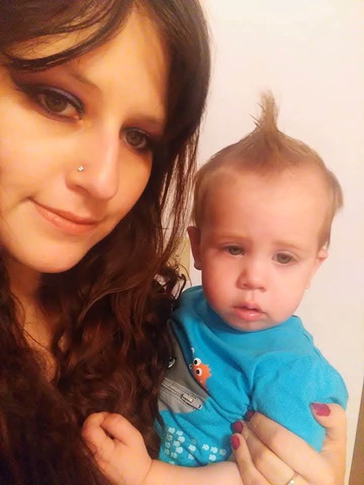 A woman with long dark hair and a nose ring holds a baby with a blue shirt and a small mohawk.