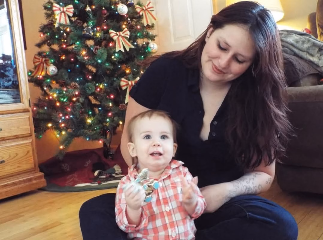 A woman with long dark hair and a baby in a plaid shirt sitting on a wooden floor in front of a decorated Christmas tree.