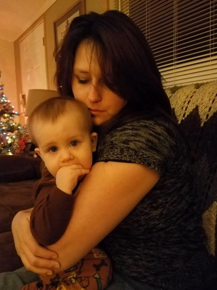 A woman with dark hair embraces a baby in a brown shirt, with a lit Christmas tree in the background.