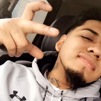 Marcos, 28, a man with a goatee and neck tattoos, makes a hand gesture while wearing a grey Under Armour hoodie.