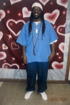 Michael, 47, smiling in a blue shirt and white beanie against a heart-patterned backdrop.