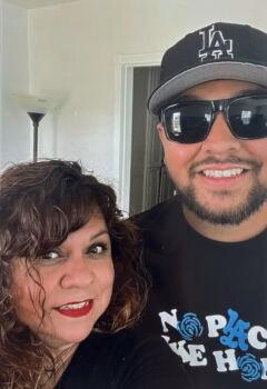 IMG_6925 A man and a woman posing for a selfie indoors. The man wears a black LA baseball cap, sunglasses, and a black t-shirt with a 'No Place Like Home' graphic. The woman has curly brown hair and red lipstick.