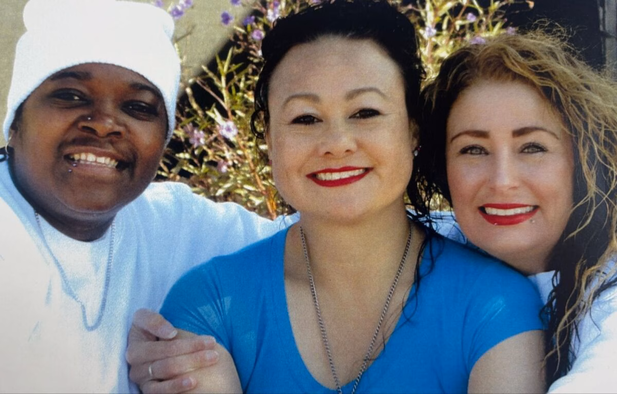 Three women smiling together outdoors, with Brenda in the center wearing a blue shirt.