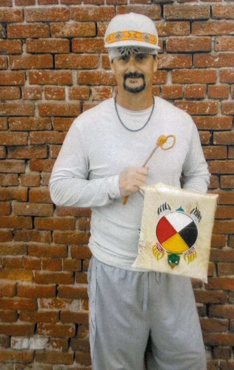 Jamie, 53, stands against a brick wall holding a bag with a colorful circular logo and a wooden stick.