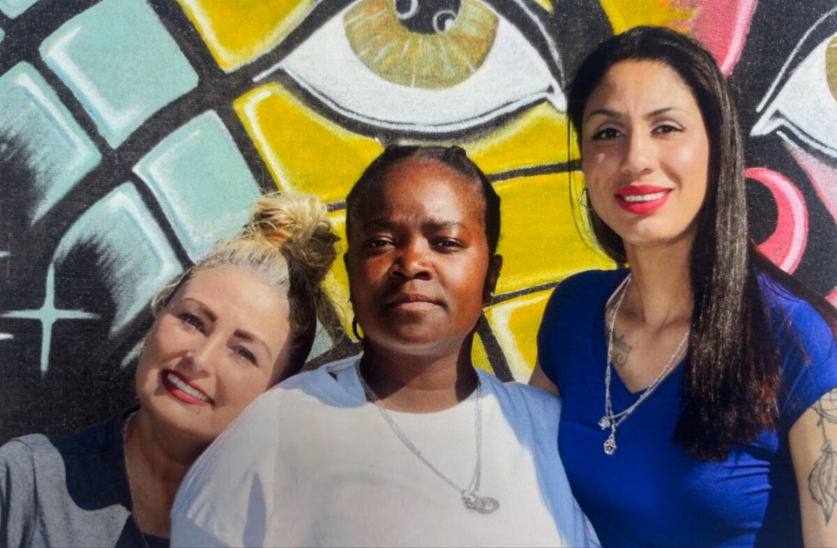Three women posing together in front of a vibrant, stylized mural featuring a large eye.