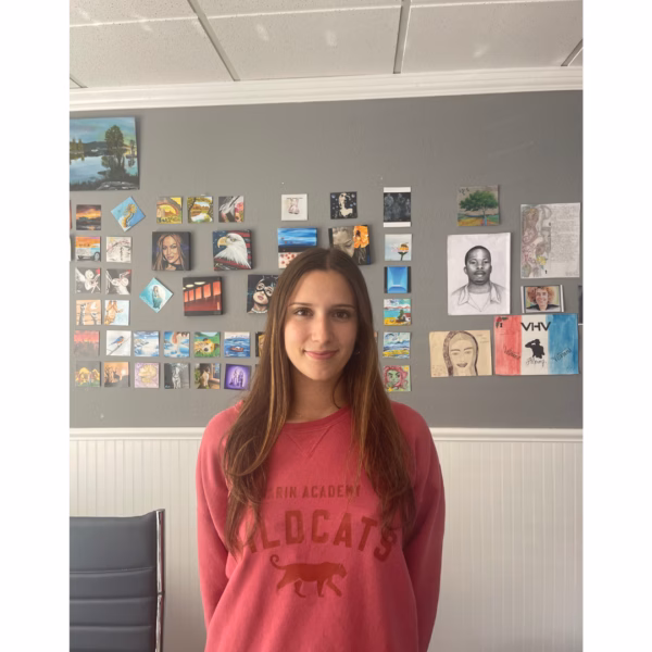 A young woman in a Cal Poly San Luis Obispo hoodie stands in front of a wall decorated with various colorful paintings and sketches.