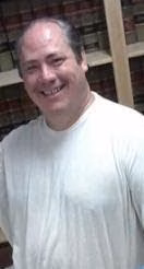 9072630031312931777 A smiling man with thinning hair wearing a white t-shirt, standing in front of a bookshelf.