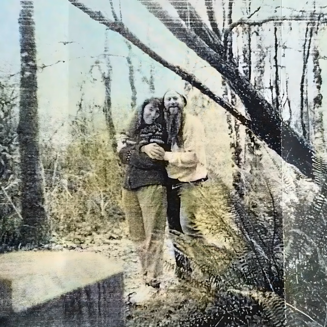 A man with a long beard and a woman stand together in a forest, framed by a large tree branch.