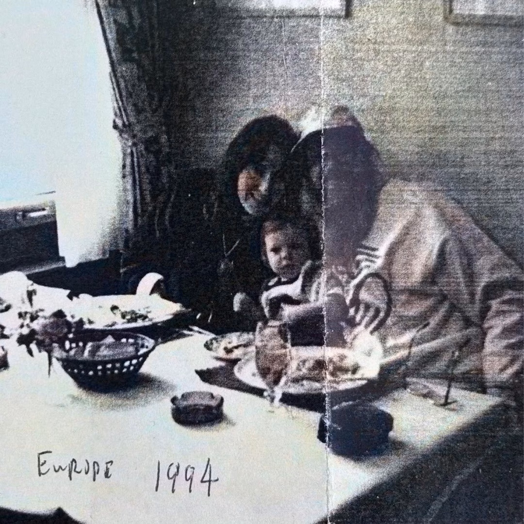 A vintage, grainy photograph of a woman and a baby at a dining table, with handwritten text 'Europe 1994' at the bottom.