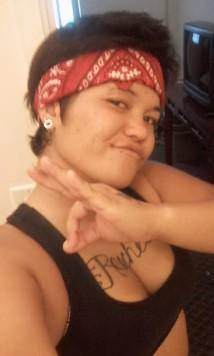 Melanie, 34, wearing a red bandana and black tank top with a 'Pinky' tattoo, making a heart shape with her hands.