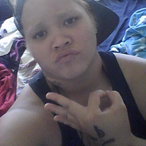Melanie, 34, a woman with a headband and a black tank top, looks at the camera while making a heart shape with her hand. A tattoo is visible on her wrist.