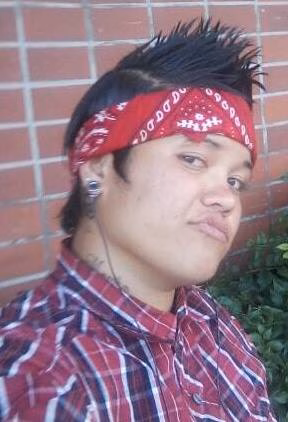 A young man with a red bandana and plaid shirt poses against a brick wall.