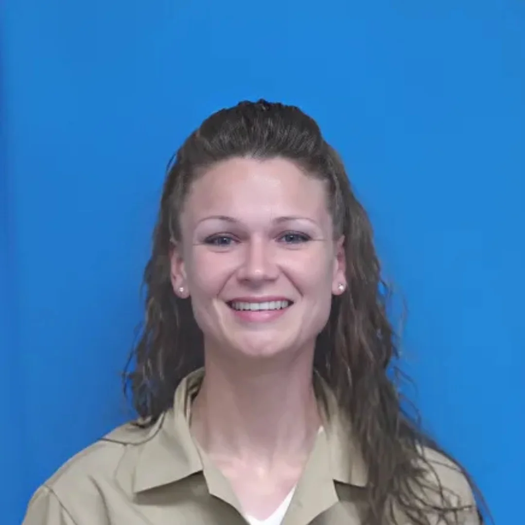 Headshot of a smiling woman with long brown hair wearing a tan collared shirt against a solid blue background