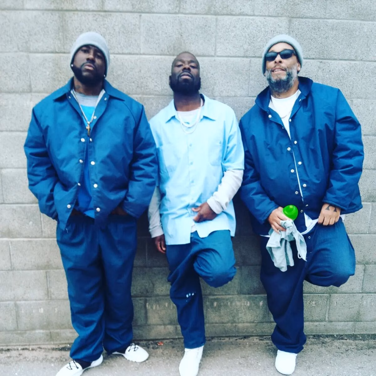 Three men in blue outfits pose against a grey brick wall.