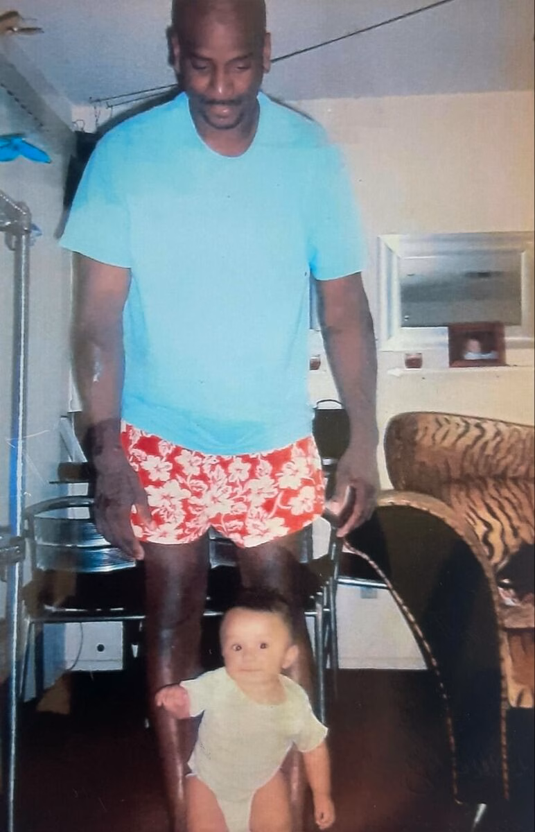 A tall man in a blue shirt and red floral shorts stands behind a baby in a white onesie in a room.