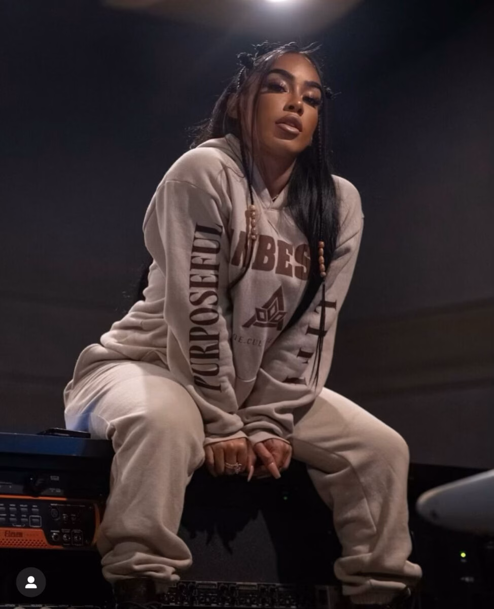 A woman with long braided hair sits on audio equipment, wearing a beige sweatshirt with 'PURPOSEFUL' and 'UMBES' text.