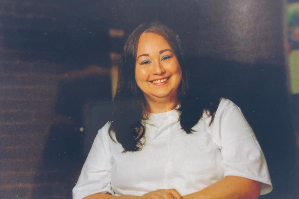 Portrait of Patricia, 55, smiling warmly in a white shirt against a dark background.