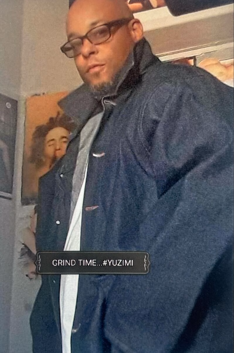 A man with glasses and a beard wearing a dark jacket over a white shirt, with a text overlay that reads 'GRIND TIME...#YUZIMI'.