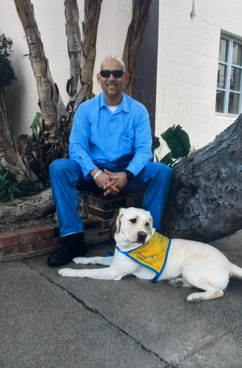 A smiling man in a blue shirt and sunglasses sits on a brick ledge next to a white service dog wearing a yellow vest that says 'Canine Companions'.