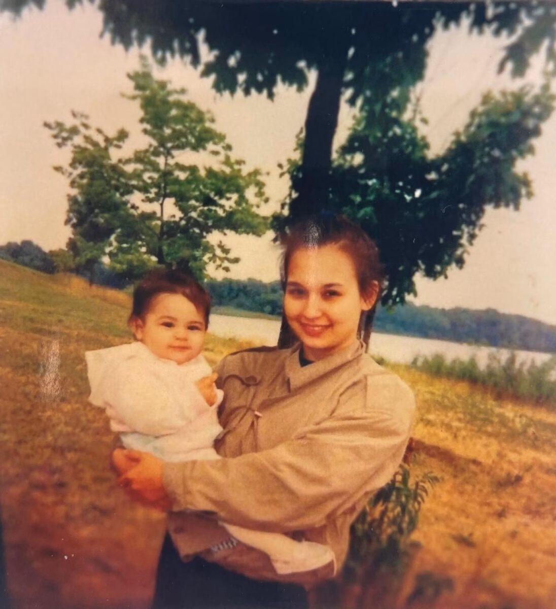A woman named Patricia holds a baby in a park with trees and a lake in the background.