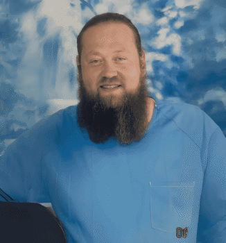 Portrait of Stewart, 47, a man with a full beard and blue eyes, wearing a light blue shirt with a pocket logo, against a blue cloudy background.