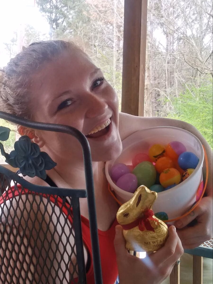 A smiling woman in a red top holds a white bucket filled with colorful plastic Easter eggs and a large gold-wrapped chocolate bunny.