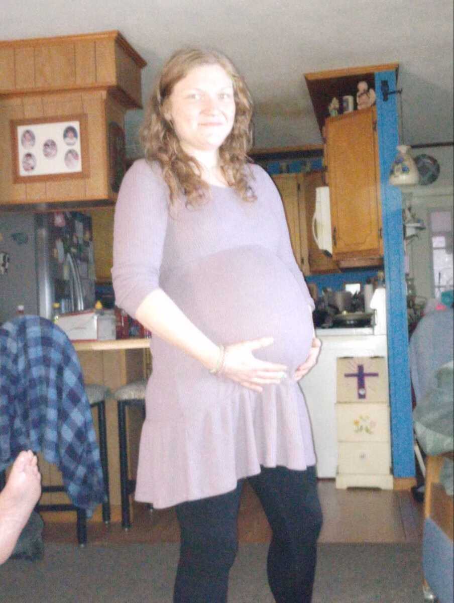 A pregnant woman with long brown hair stands in a kitchen, wearing a purple top and black leggings, with her hands resting on her belly.