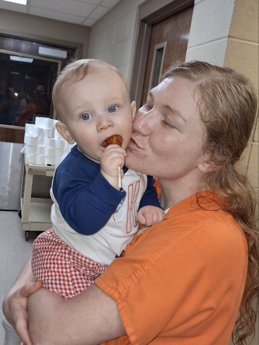 A woman in an orange shirt kisses a baby's cheek while the baby eats a lollipop.