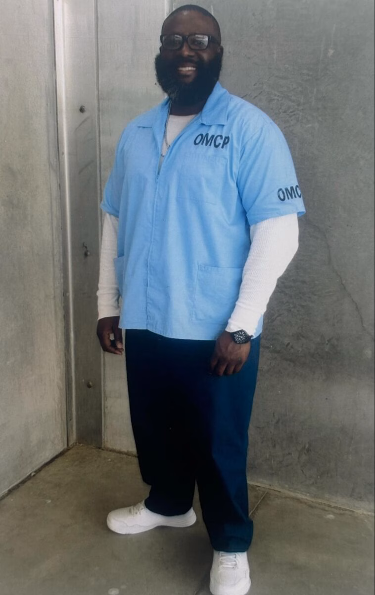 A smiling Black man with a beard and glasses wearing a light blue OMCP uniform shirt over a white long-sleeved shirt, dark blue pants, and white sneakers.