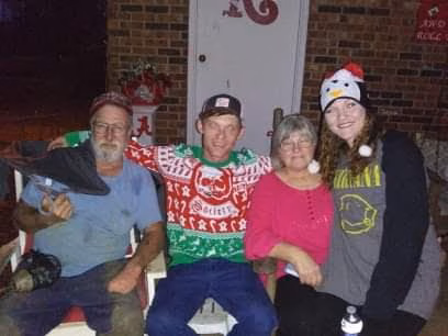 Four people sitting together in front of a brick wall, wearing festive holiday-themed clothing.