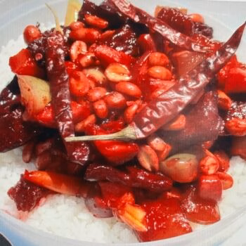 A rice bowl topped with a vibrant red sauce, featuring whole peanuts, sliced garlic, and several long, dried red chili peppers.