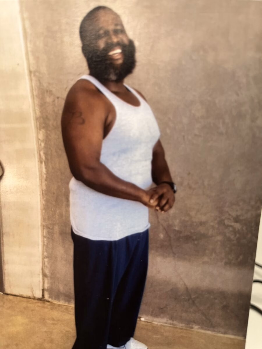 A man with a beard and a white tank top stands against a textured wall, smiling.