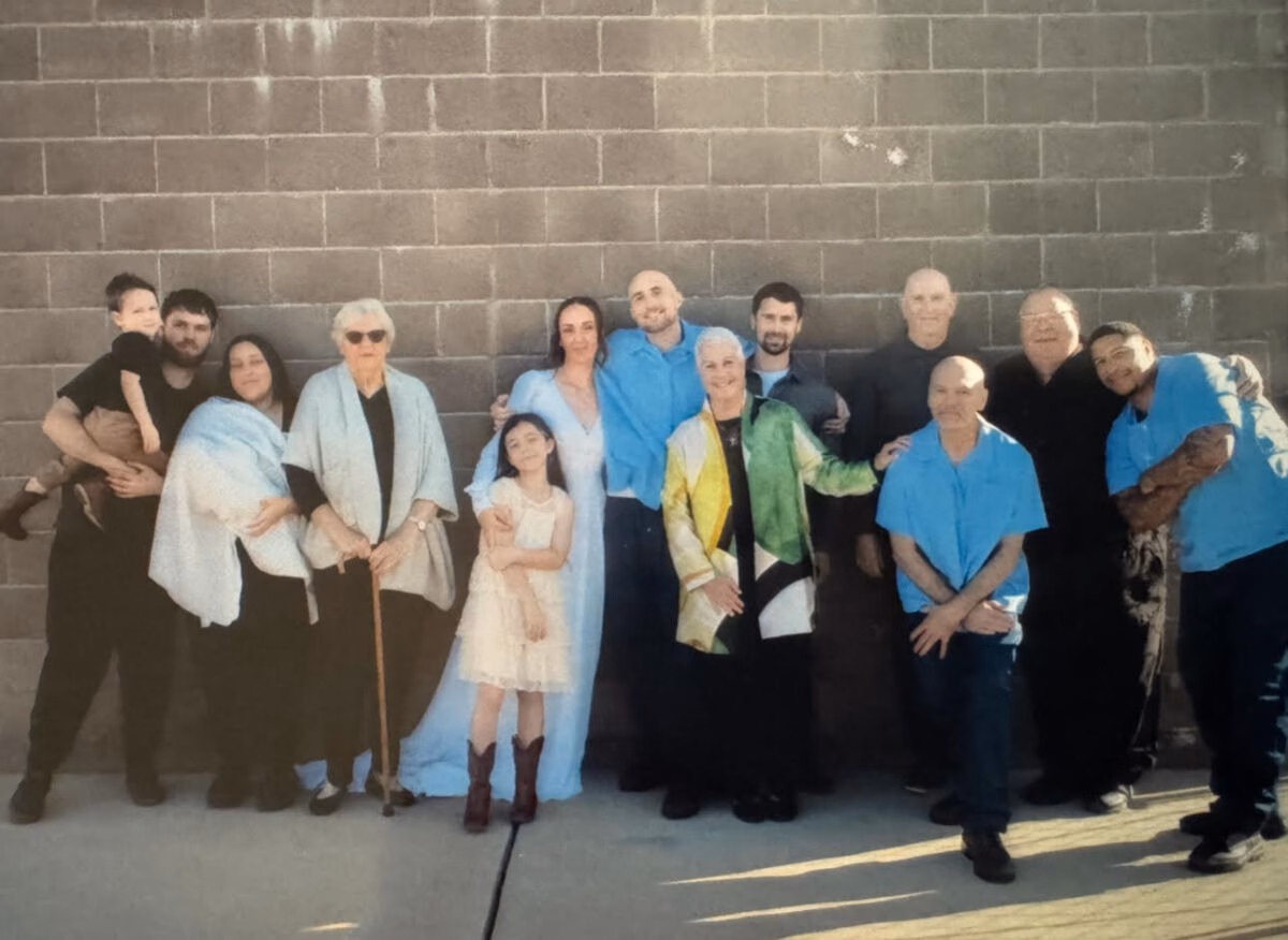 A group of thirteen people, including men, women, and children of various ages, posing together in front of a grey brick wall.