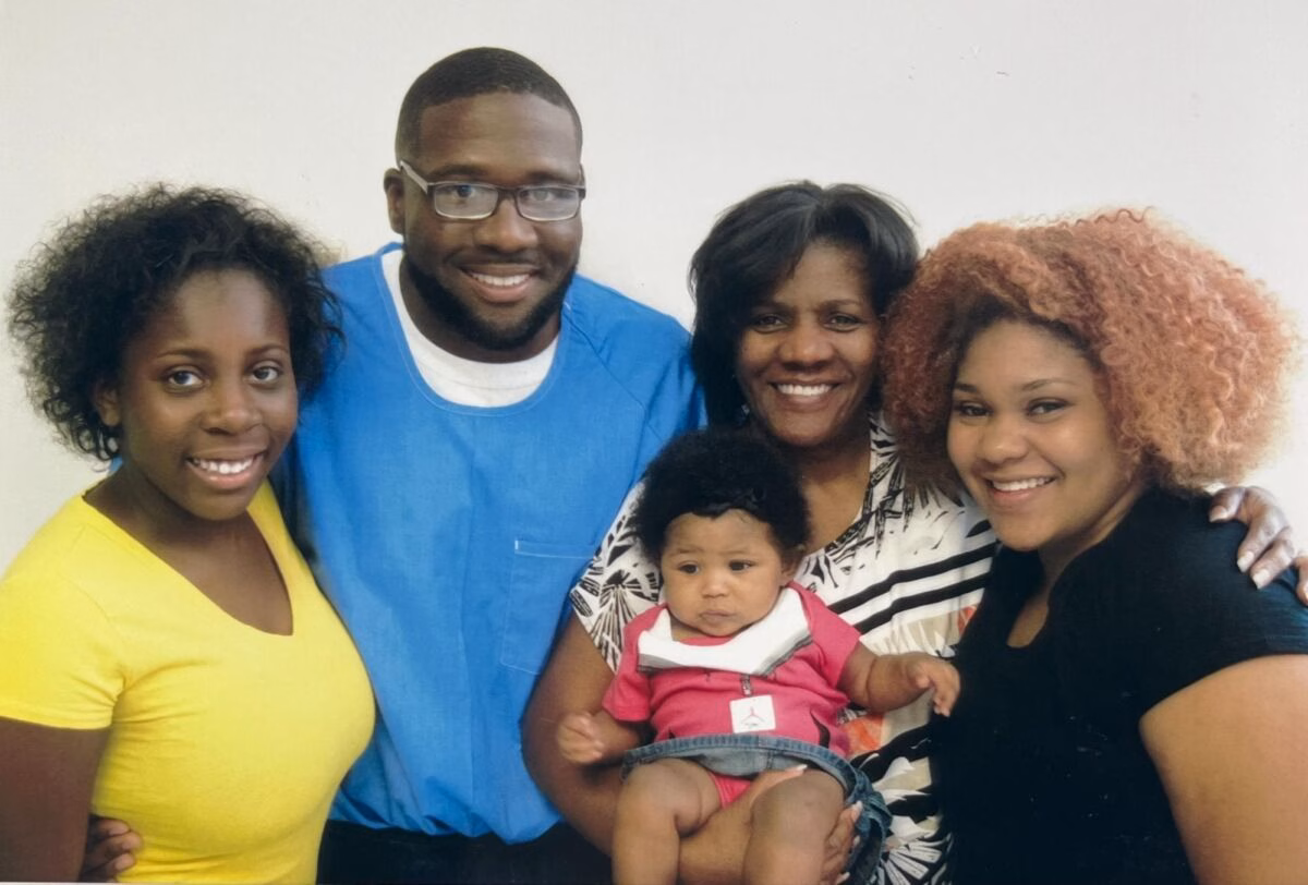 A smiling family of five, including a man in blue scrubs, two women, and a baby, posing together against a plain wall.