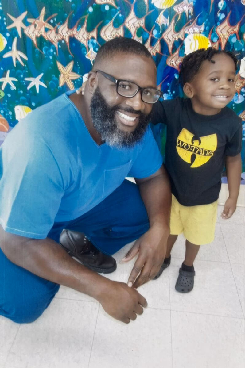 A smiling man with a beard and glasses in a blue shirt kneels next to a young boy in a Wu-Tang Clan t-shirt, both in front of a colorful mural.