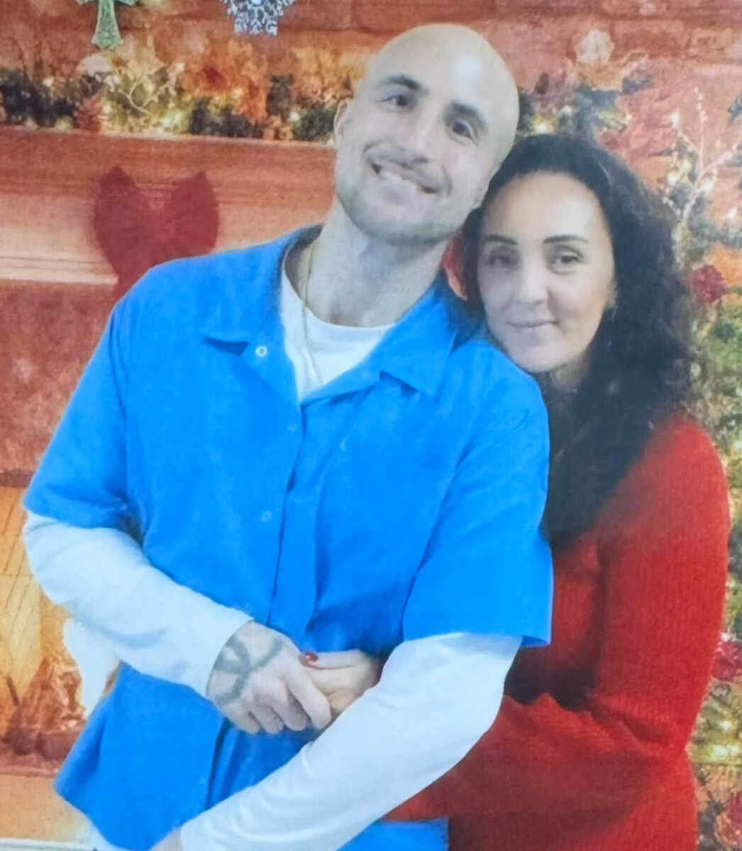 A bald man in a blue shirt and a woman in a red sweater standing together in front of a fireplace.