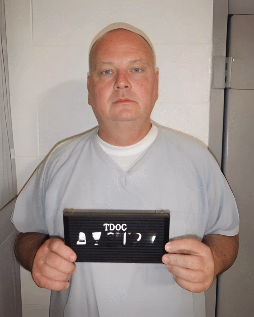 A man in a light blue prison uniform and a white cap holds a black mugshot board with the name 'TDOC' and several symbols.