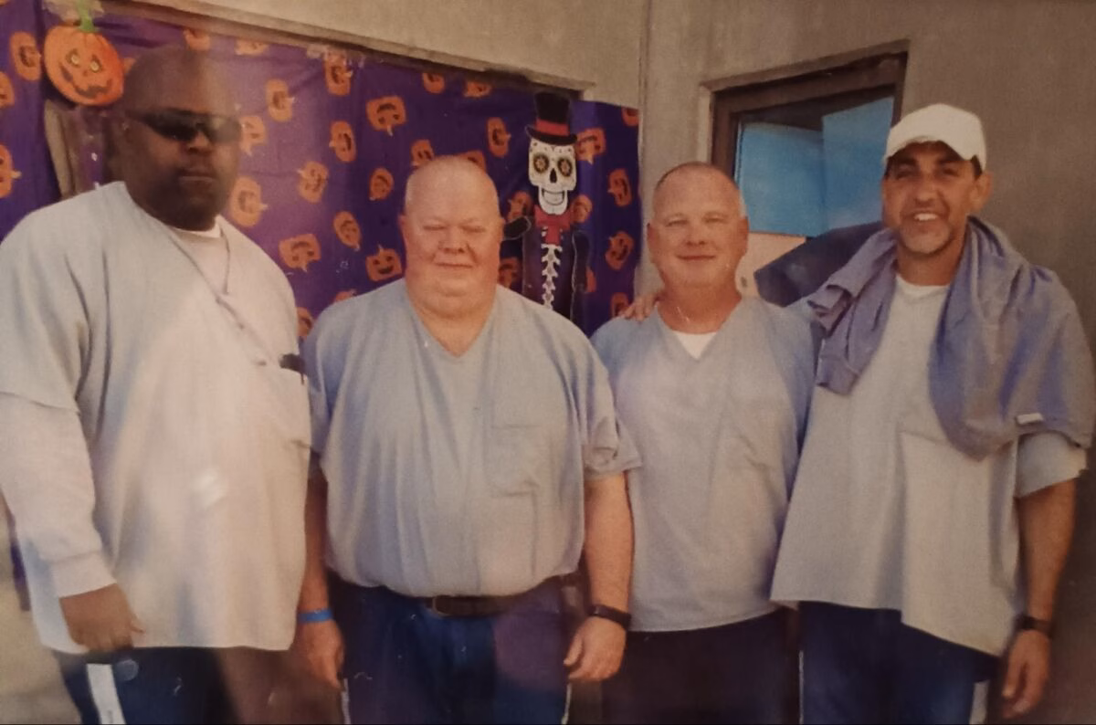 Four men in light blue shirts stand together in front of a purple Halloween-themed backdrop.