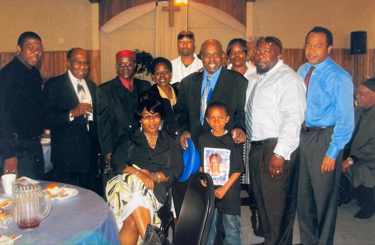 A group of eleven people, including a young boy in a t-shirt, posing for a photo in a church hall with a cross on the wall.