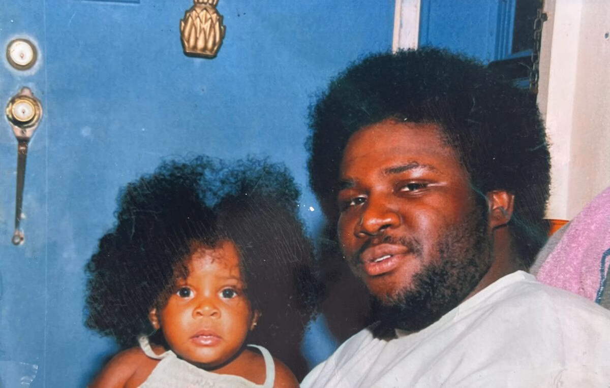 A man and a young child with large, curly hair stand in front of a blue door with a pineapple-shaped knocker.
