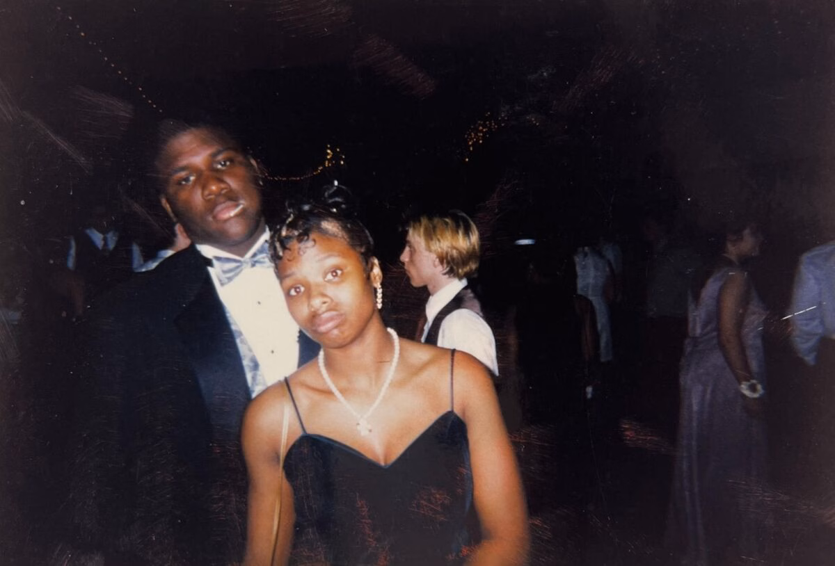 A young man and woman in formal wear pose together at a dimly lit event, with other guests visible in the background.