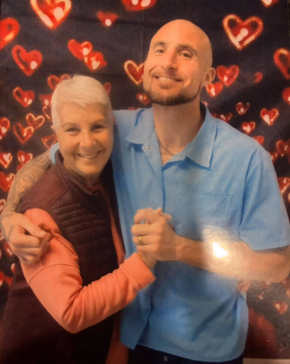 Anthony, 38, smiling with an older woman against a backdrop of red hearts.