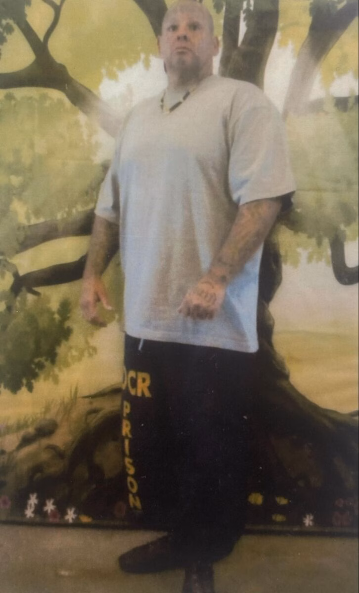 A man with tattoos on his arms and hands stands in front of a mural of a large tree. He is wearing a light-colored t-shirt and black pants with the words 'OCR PRISON' printed vertically in yellow.