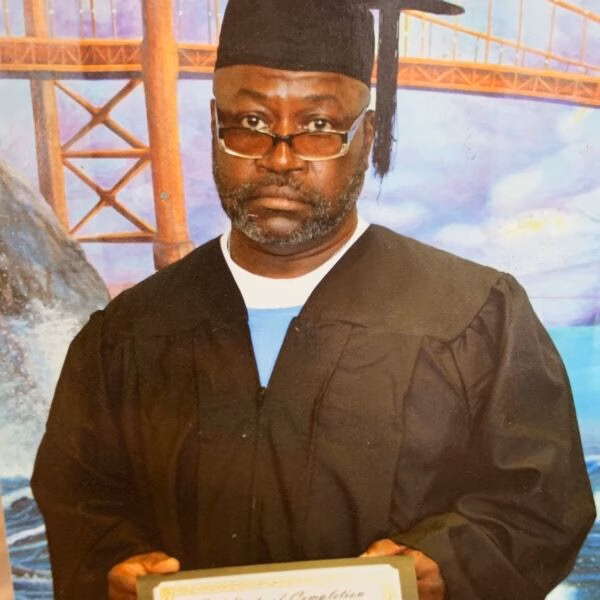 Rodney Baylis in a black graduation cap and gown, holding a certificate of completion for the Vocational Computer Literacy Class, with the Golden Gate Bridge in the background.