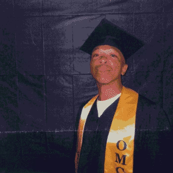 Kenneth, 43, in a black graduation cap and gown with a yellow sash that reads OMC, smiling against a dark backdrop.