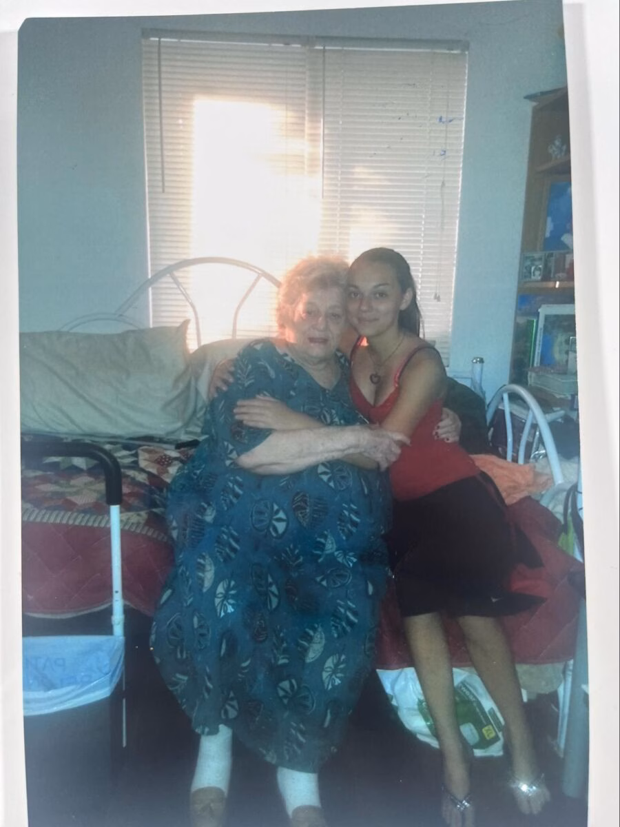 An elderly woman in a patterned blue dress sits on a bed, embracing a young woman in a red top and black skirt.