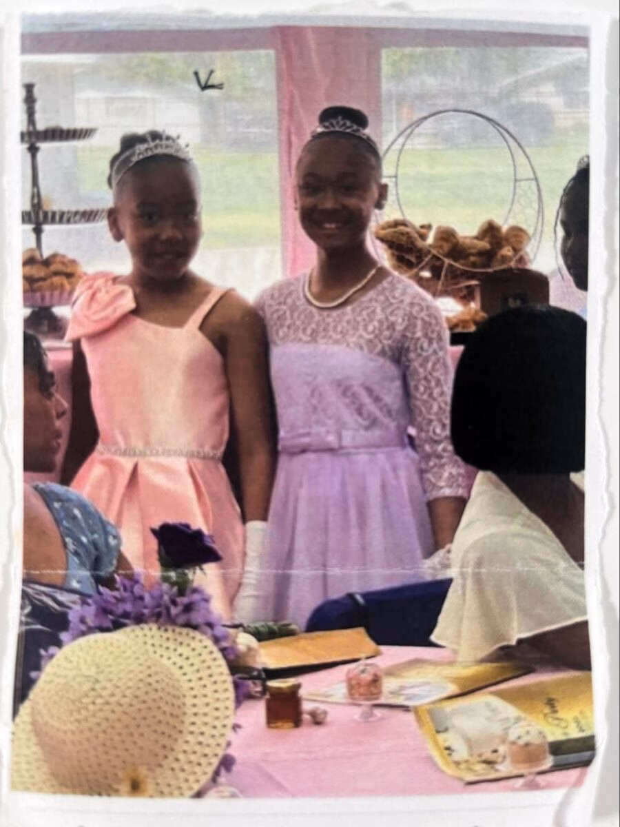 image13 Two young girls in formal dresses and tiaras stand together at a table with a pink tablecloth, surrounded by other people at an event.