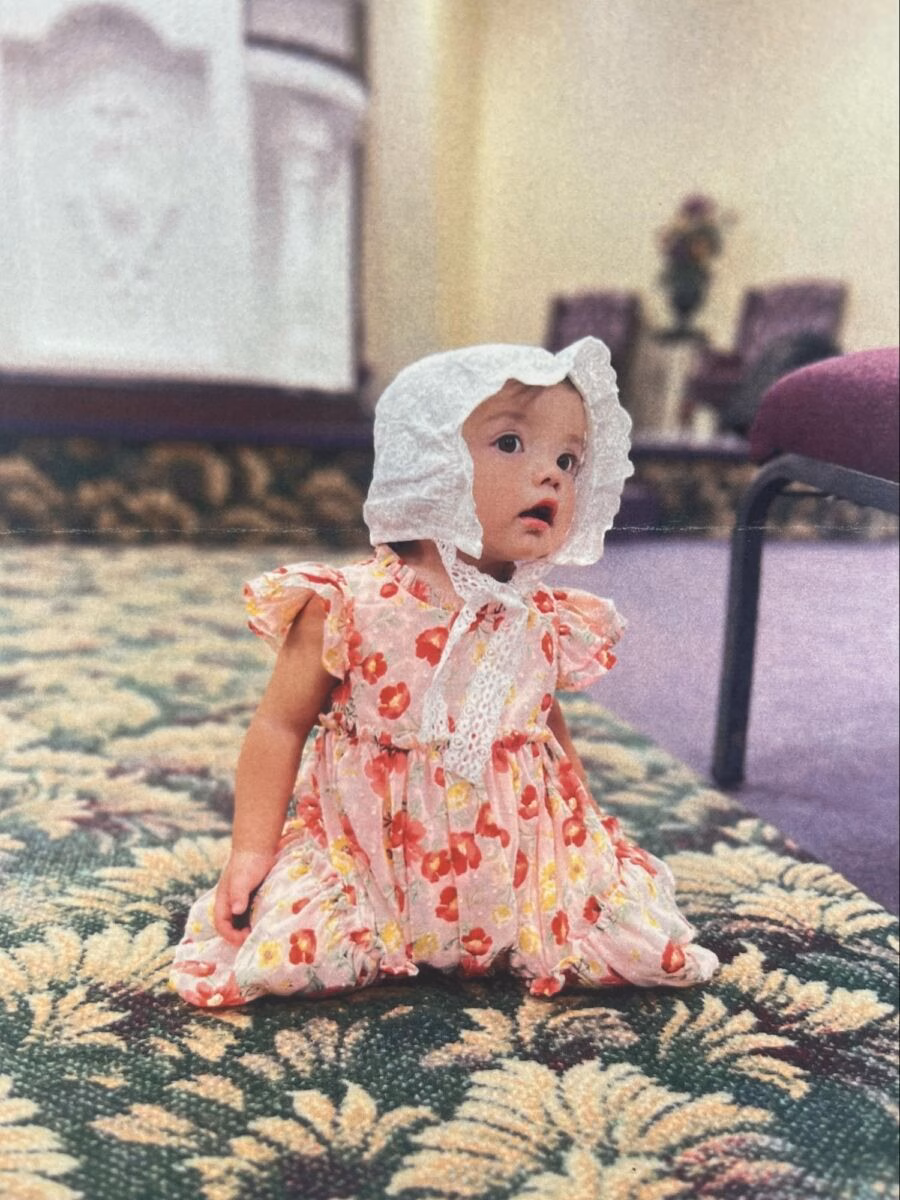 A young baby wearing a white bonnet and a pink floral dress sits on a patterned carpet.