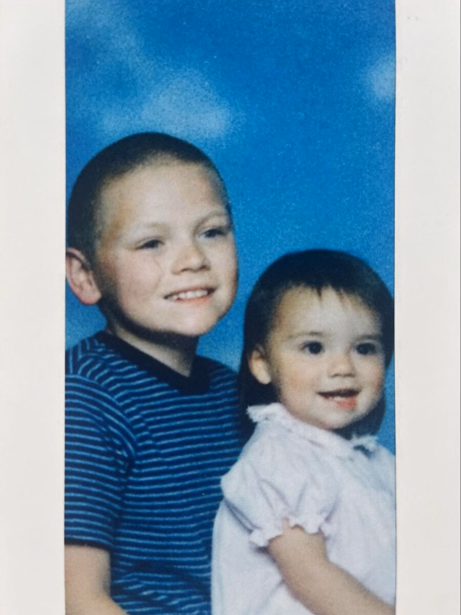 Portrait of David, 33, a young boy with a shaved head in a striped shirt, next to a smiling toddler in a white dress against a blue background.